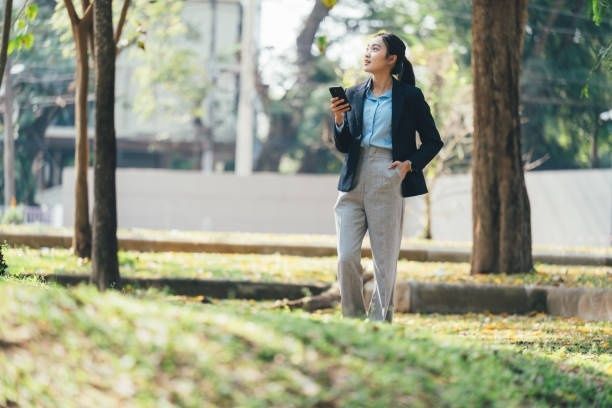 Affärsklädd kvinna promenerar med mobilen i en grön stadspark